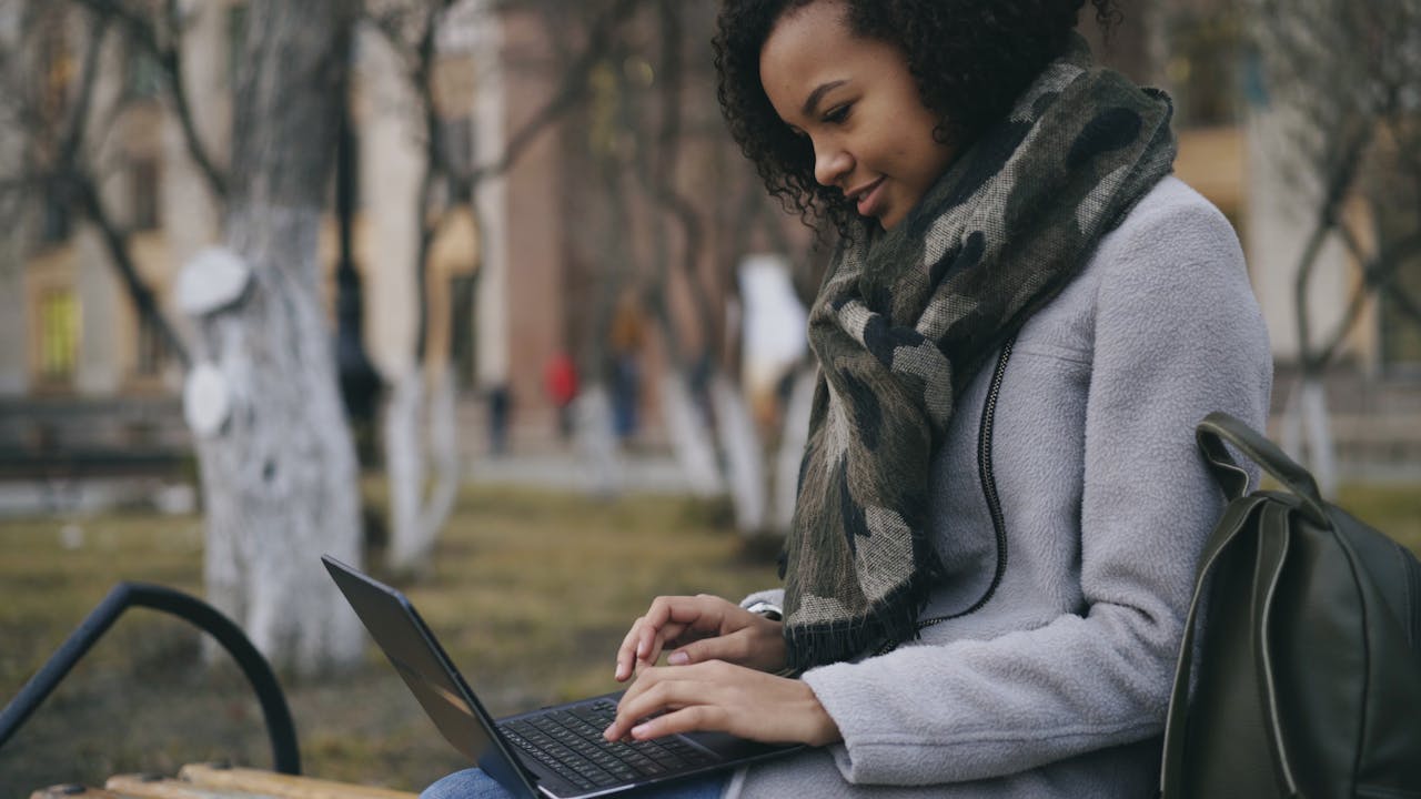 A young woman sits outside using her laptop, bundled up in a scarf.